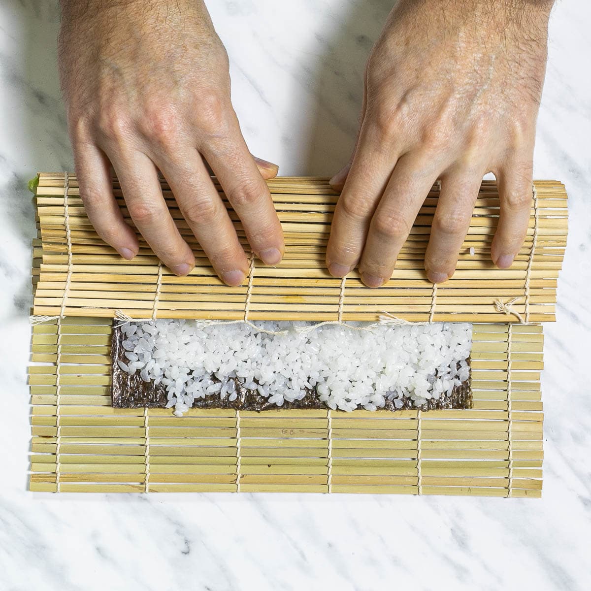A bamboo mat with seaweed sheet and rice. A hand is rolling it so far until half is covered. 