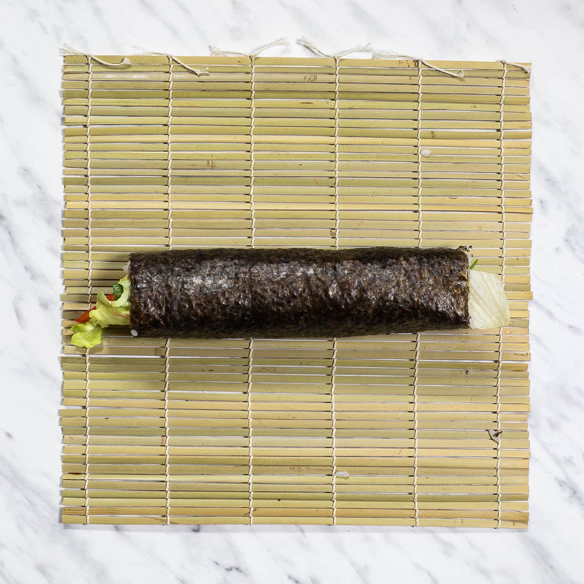 A bamboo mat with a long rolled sushi covered in seaweed sheets. Some of the fillings can be seen at the ends. 