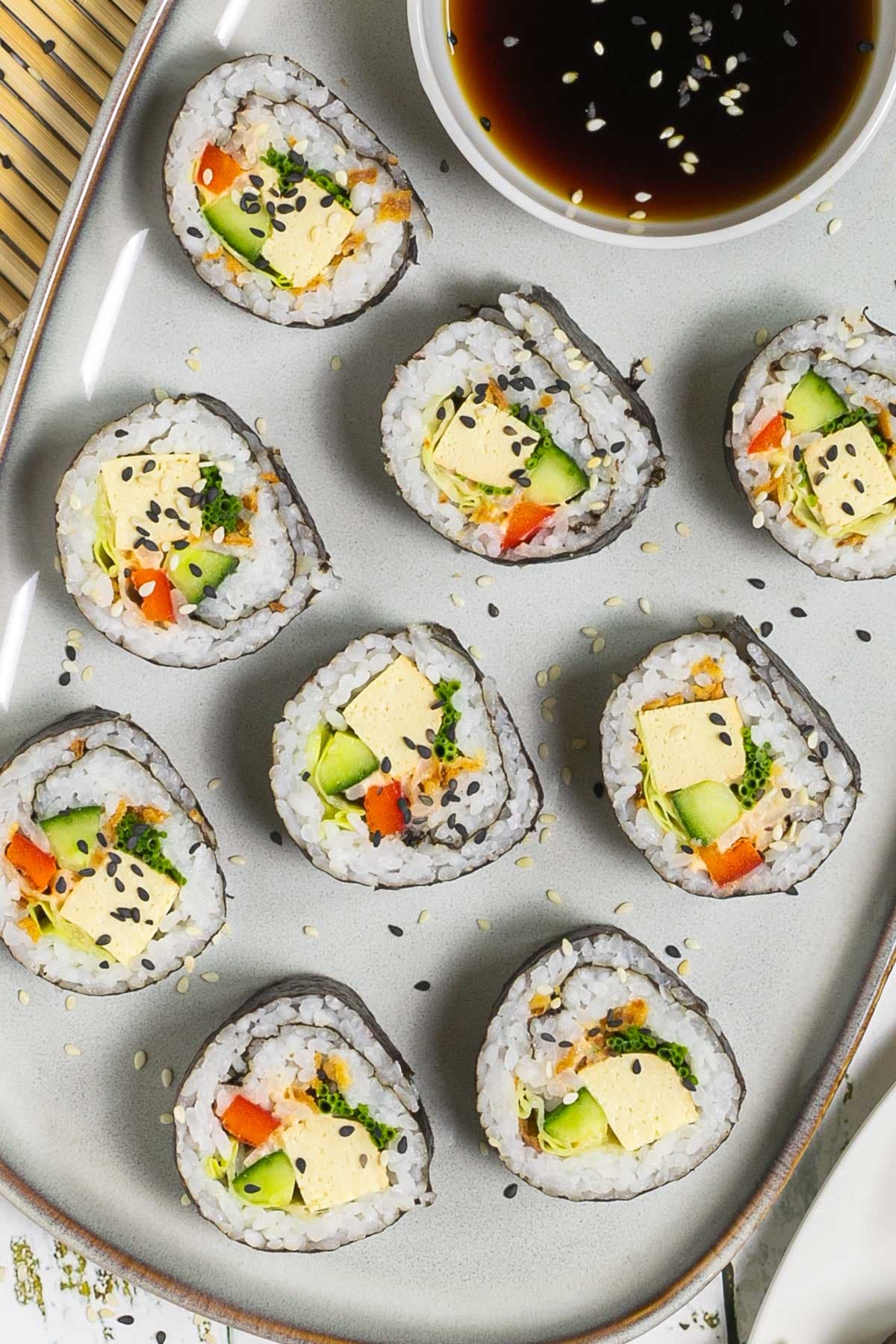 An oval, light blue plate with 9 tofu sushi rolls with colorful filling wrapped in dark seaweed sheets. Served with a small bowl of soy sauce and sprinkled with dark and white sesame seeds. 