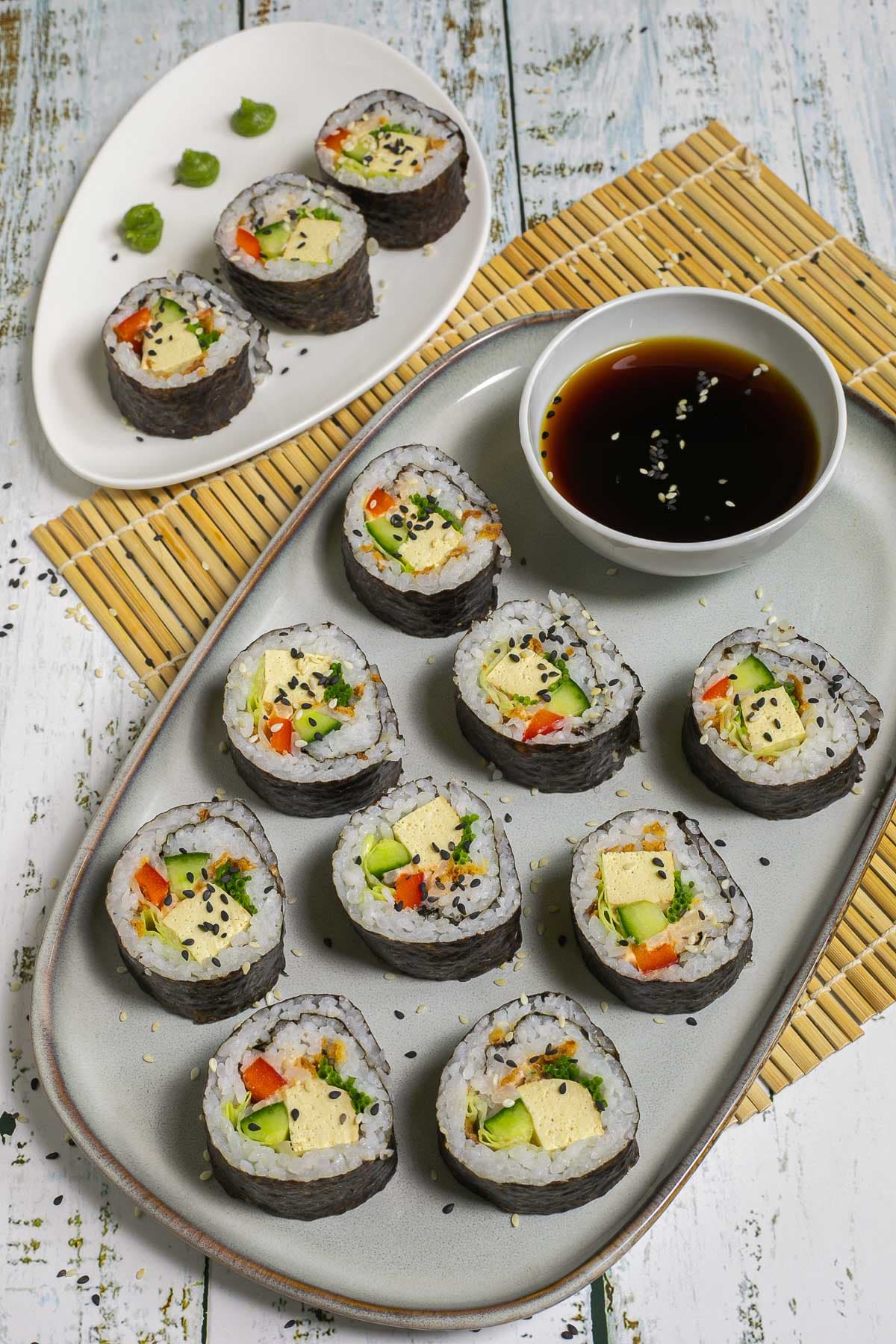 An oval, light blue plate with 9 tofu sushi rolls with colorful filling wrapped in dark seaweed sheets. Served with a small bowl of soy sauce and sprinkled with dark and white sesame seeds.