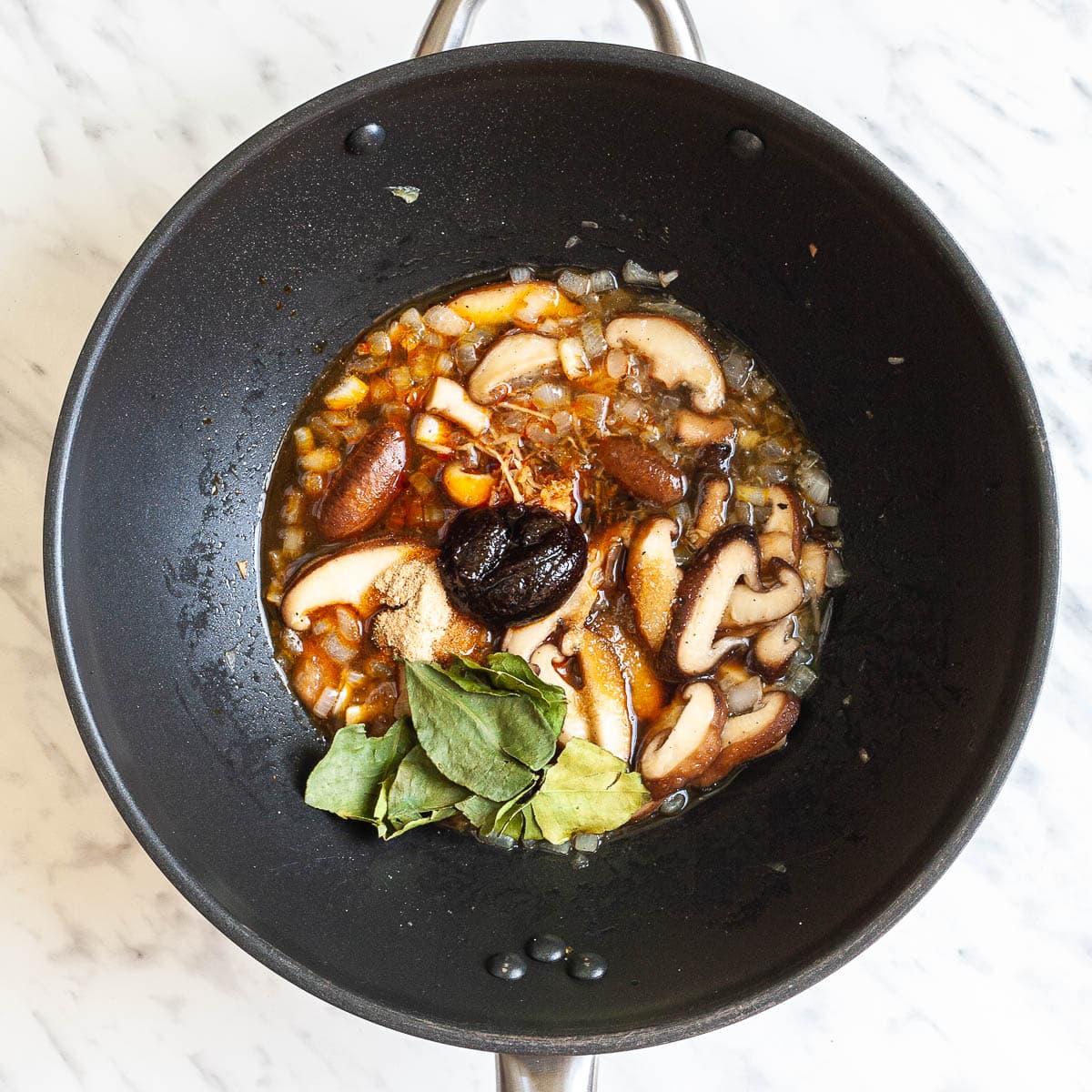 Black wok with mushroom slices, green leaves, dark brown paste and different orange and brown spices