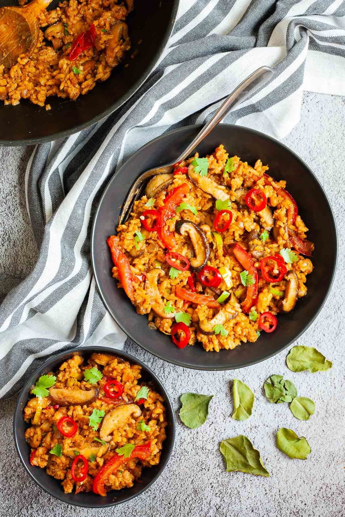 2 black bowls with orange brown fried rice, sliced mushrooms, slices tomatoes, chopped cilantro and sliced chili pepper. A wok with the leftovers is in the corner. Kaffir lime leaves are placed around in a grey surface.
