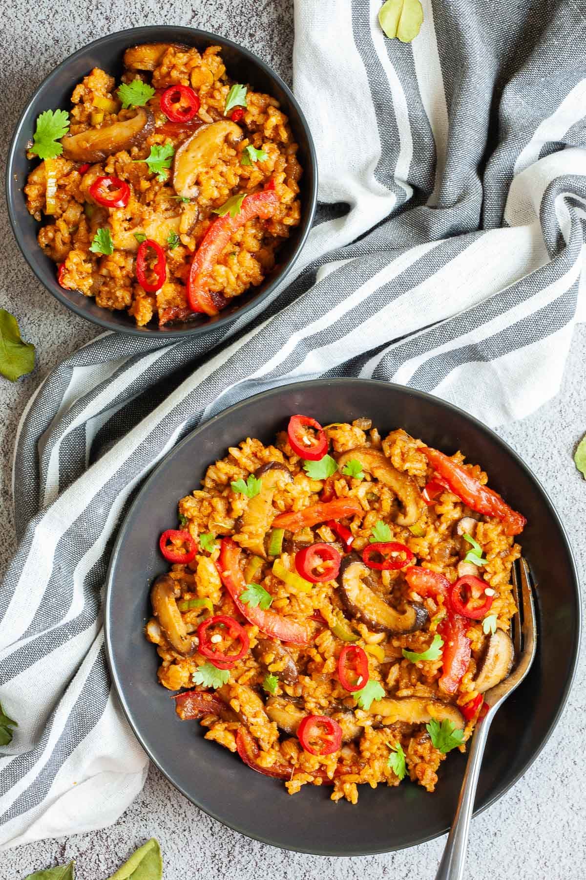 2 black bowls with orange brown fried rice, sliced mushrooms, slices tomatoes, chopped cilantro and sliced chili pepper. A fork is placed in one of them.