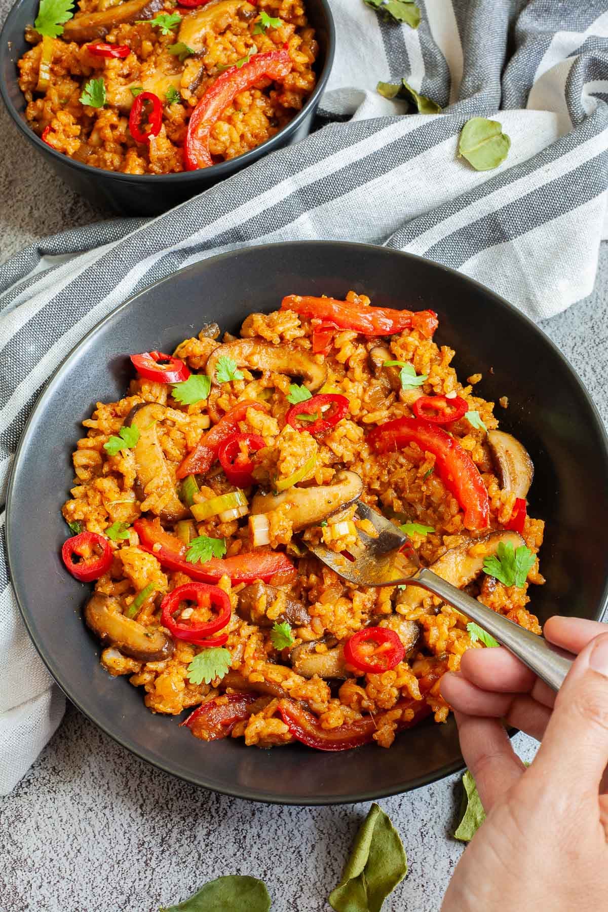 2 black bowls with orange brown fried rice, sliced mushrooms, slices tomatoes, chopped cilantro and sliced chili pepper. A hand is holding a fork to take a bite.