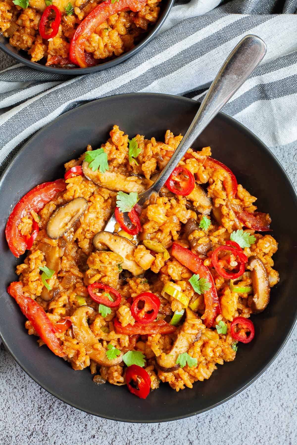 2 black bowls with orange brown fried rice, sliced mushrooms, slices tomatoes, chopped cilantro and sliced chili pepper. A fork is placed inside.