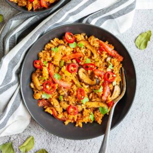 A black bowl with orange brown fried rice, sliced mushrooms, slices tomatoes, chopped cilantro and sliced chili pepper. A fork is placed in one of them.