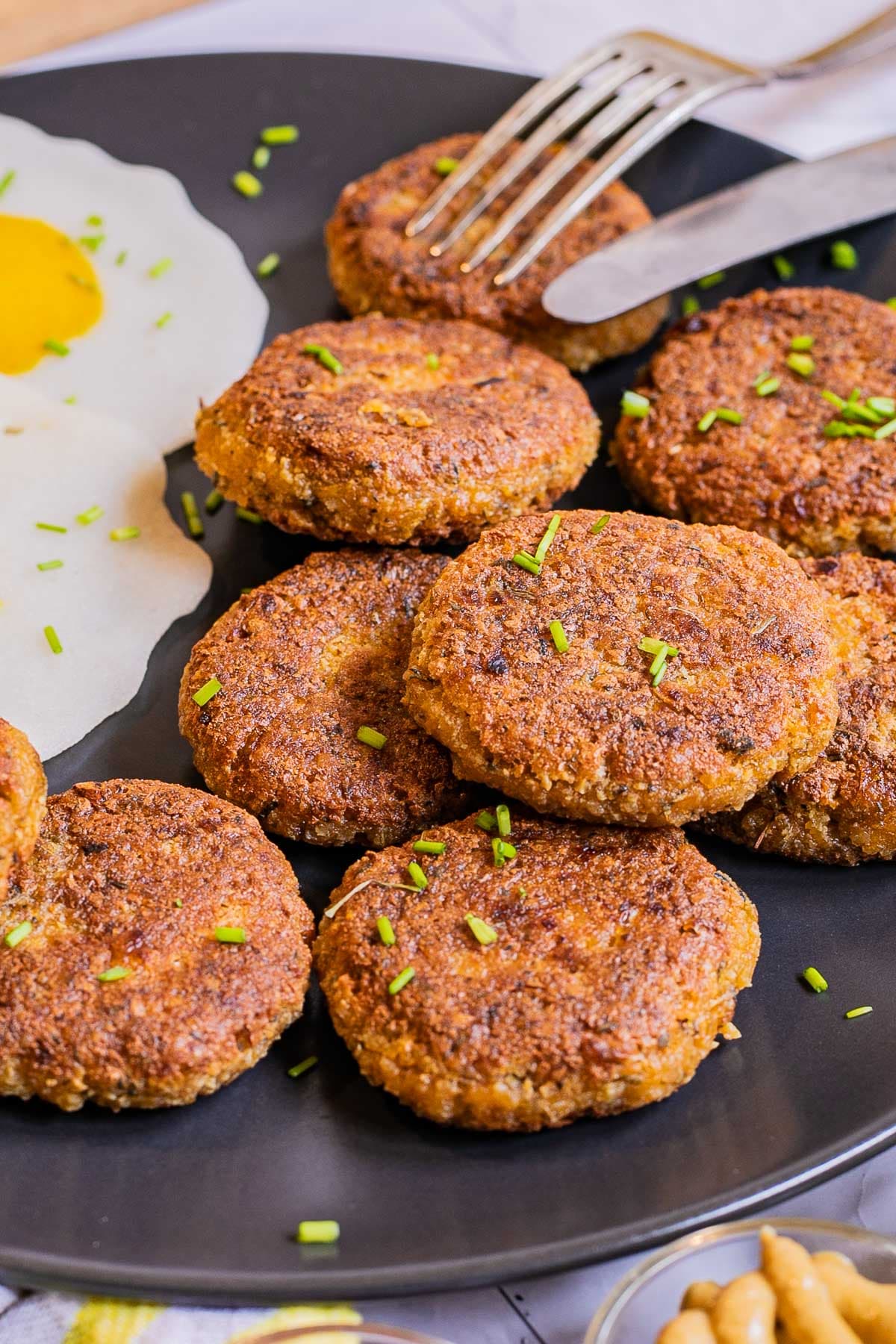 Nine round flat crispy brown sausage patties on a black plate served with fried egg look-alikes. Utensils are placed on top.