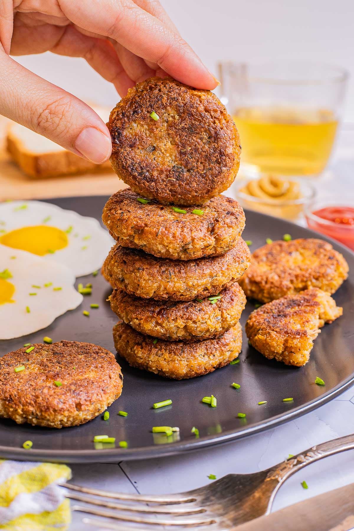 A stack of round flat crispy brown sausage patties on a black plate served with fried egg look-alikes. A hand is taking the top one.