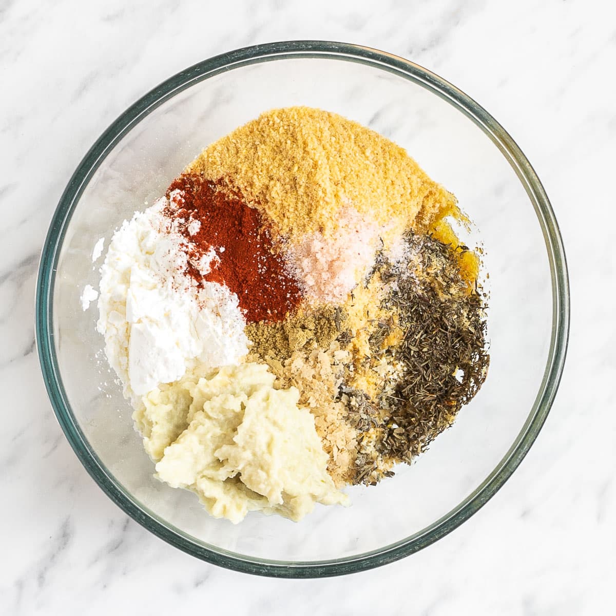 All ingredients for vegan breakfast sausage is in a glass bowl before mixing them. All ingredients are visible, tofu mince, breadcrumbs, starch, different spices and onion-garlic puree.
