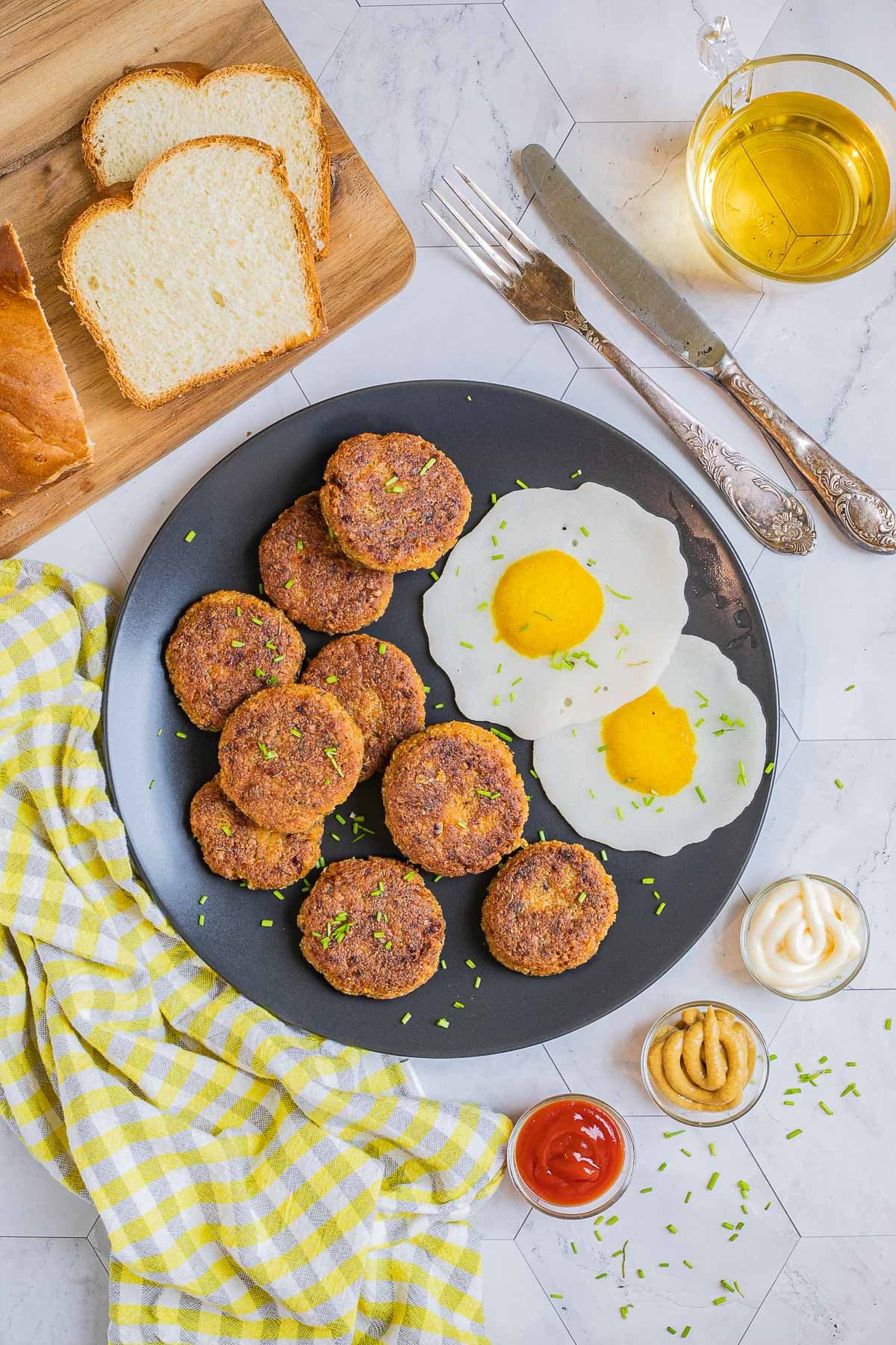 Nine round flat crispy brown sausage patties on a black plate served with fried egg look-alikes. Condiments, bread, utensils, and a glass of juice is placed next to it.