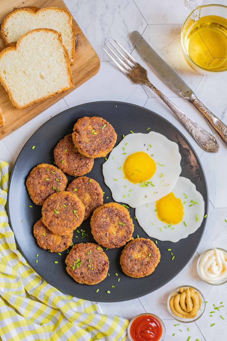 Nine round flat crispy brown sausage patties on a black plate served with fried egg look-alikes. Condiments, bread, utensils, and a glass of juice is placed next to it.
