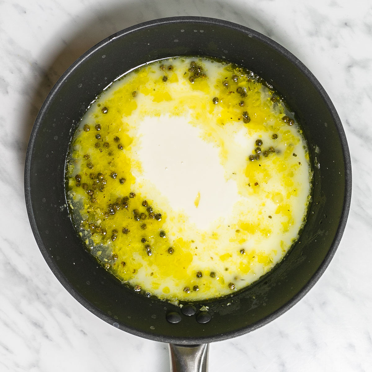 Black frying pan with a yellowish sauce and whole peppercorn, in the middle a white creamy sauce is added.