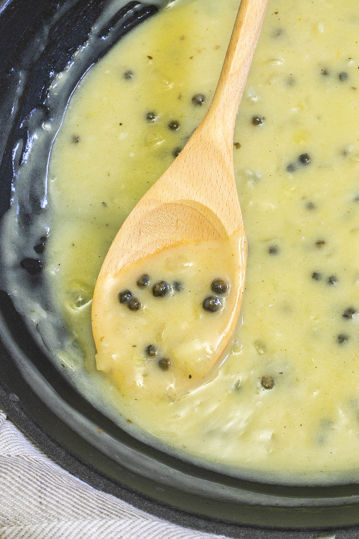 Black frying pan with a light green sauce with whole peppercorns. A wooden spoon is placed in the sauce covered by some of it.