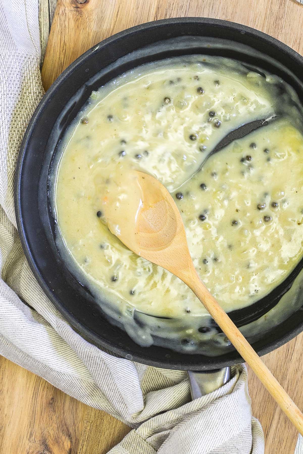 Black frying pan with a light green sauce with whole peppercorns. A wooden spoon is placed in the middle leaving a streak in the sauce.