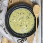 Black frying pan with a light green sauce with whole peppercorns. A wooden spoon is placed next to it.