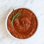 Red pesto with rosemary leaves in a small white bowl.
