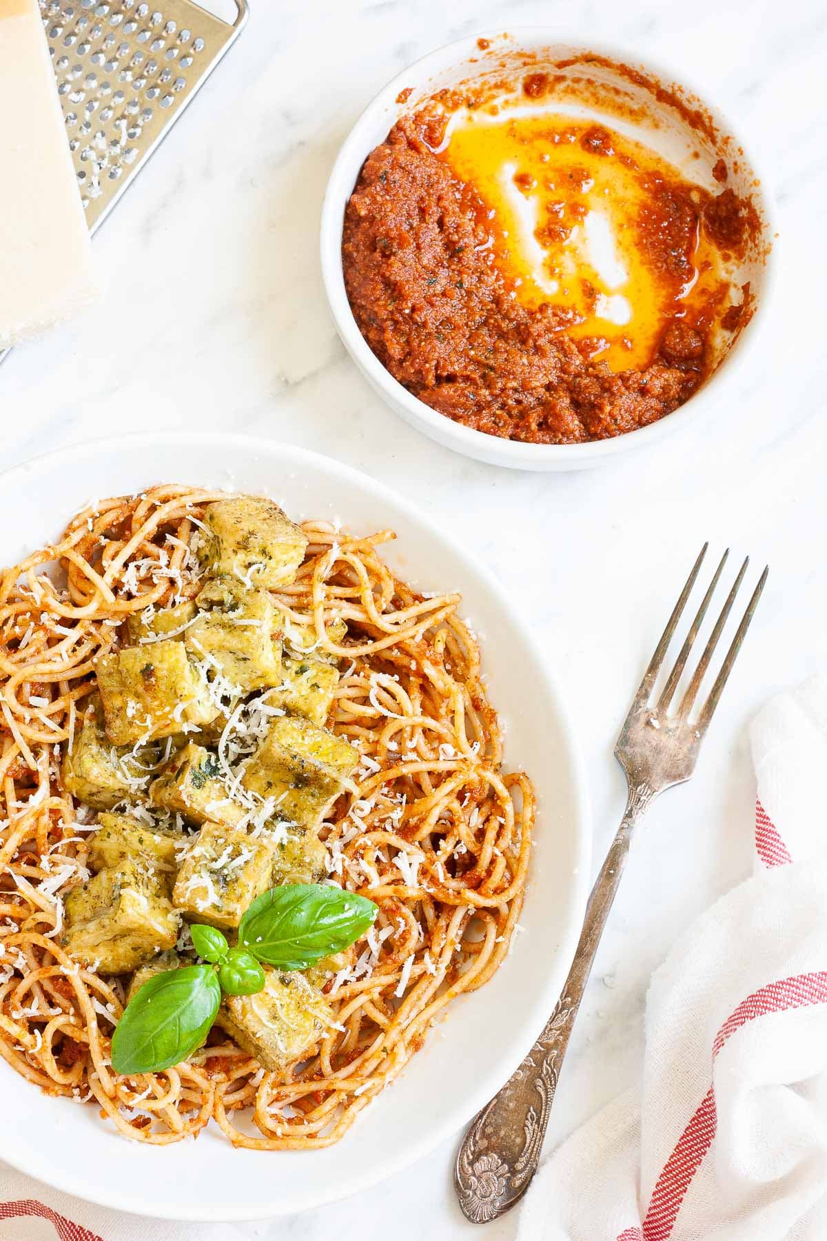 Large white plate with red pesto spaghetti topped with green-yellow tofu cubes, shredded cheese and basil leaves. There is a small bowl with leftover red pesto in the corner and parmesan cheese on top of a grater.