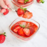2 small glass bowls with white pudding with pearls topped with strawberry sauce and fresh strawberry slices. A hand is holding a spoon with a mouthful of the pudding.