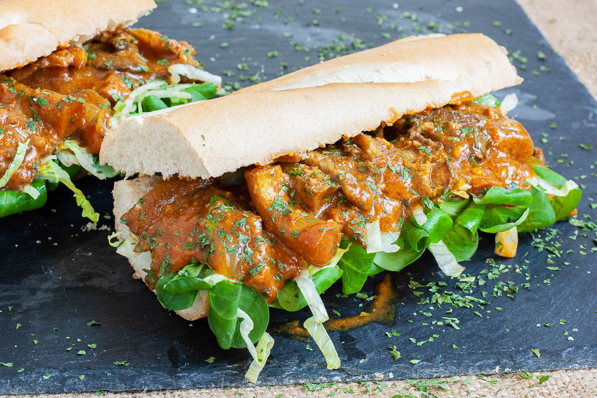 2 hoagie rolls with chopped lettuce and arugula topped with mushroom strips in a brown orange juicy sauce