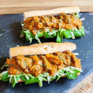 2 hoagie rolls with chopped lettuce and arugula topped with mushroom strips in a brown orange juicy sauce