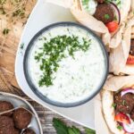 Black bowl with white green sauce topped with freshly chopped dill. The bowl is surrounded by falafel in pita bread on a white plate.