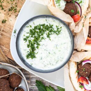 Black bowl with white green sauce topped with freshly chopped dill. The bowl is surrounded by falafel in pita bread on a white plate.