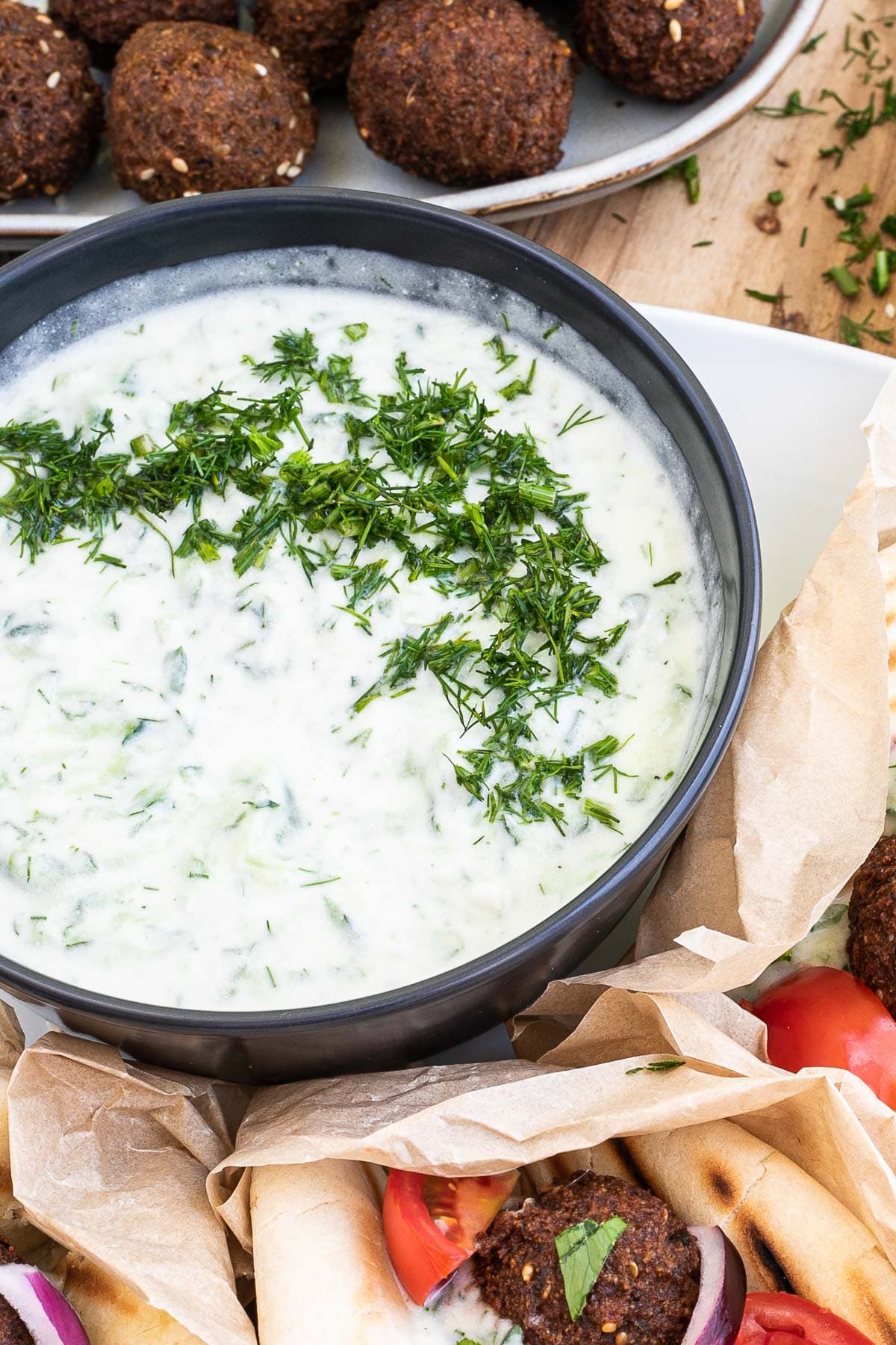 Black bowl with white green sauce topped with freshly chopped dill. The bowl is surrounded by falafel in pita bread on a white plate.