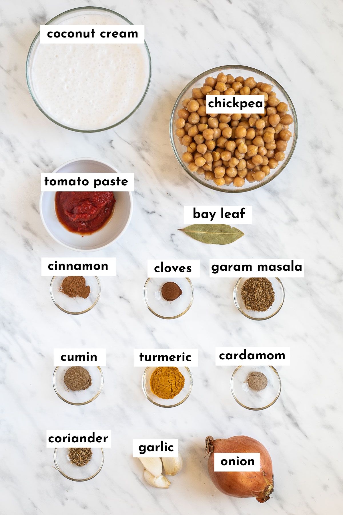 The ingredients of chickpea korma is placed in small glass bowls like coconut cream, chickpeas, tomato paste, bay leaf, and spices in different shades of brown and red.