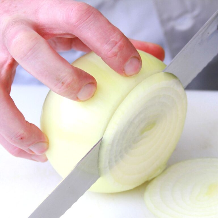 A hand is holding a large peeled onion and cutting it into slices with a large knife.