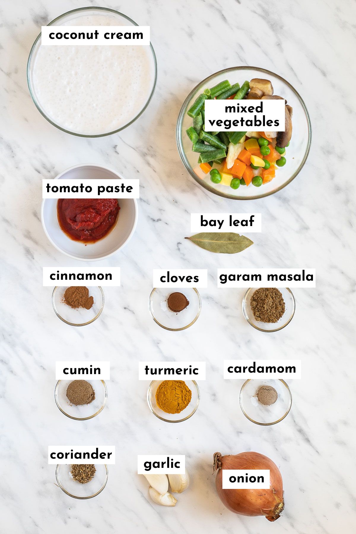 ingredients of vegetable korma in small glass bowls like white coconut milk, mixed vegetables, tomato paste, onion, garlic, bay leaves, and lots of different spices
