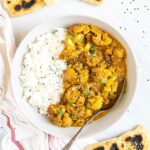 A white plate with white rice and cauliflower florets in a creamy orange sauce sprinkled freshly chopped cilantro. A spoon is placed inside. Naan bread pieces are next to it.