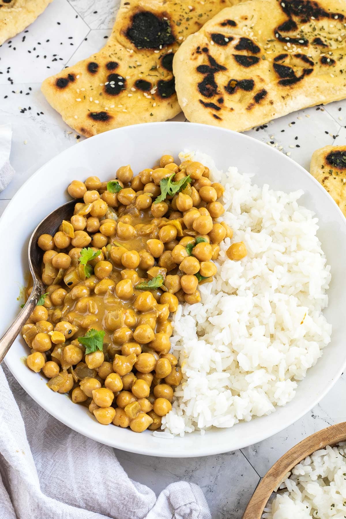 A white plate with white rice and chickpeas in a creamy orange sauce sprinkled freshly chopped cilantro. A spoon is placed inside. Naan bread and a bowl of rice are next to it.