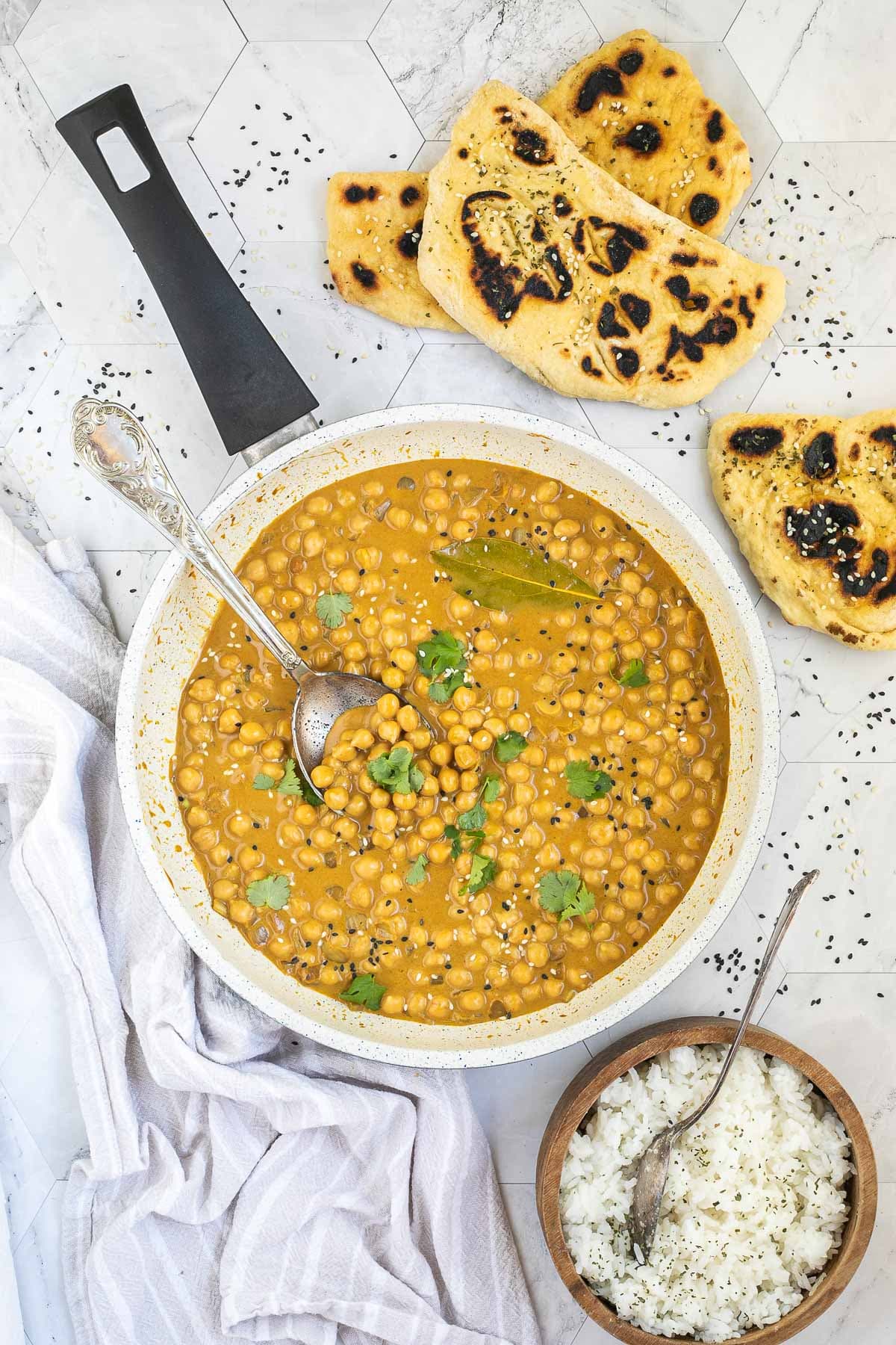 White frying pan with a creamy orange sauce mixed with chickpeas, sprinkled with sesame seeds and freshly chopped cilantro. Charred flatbread and a bowl of rice is next to it.