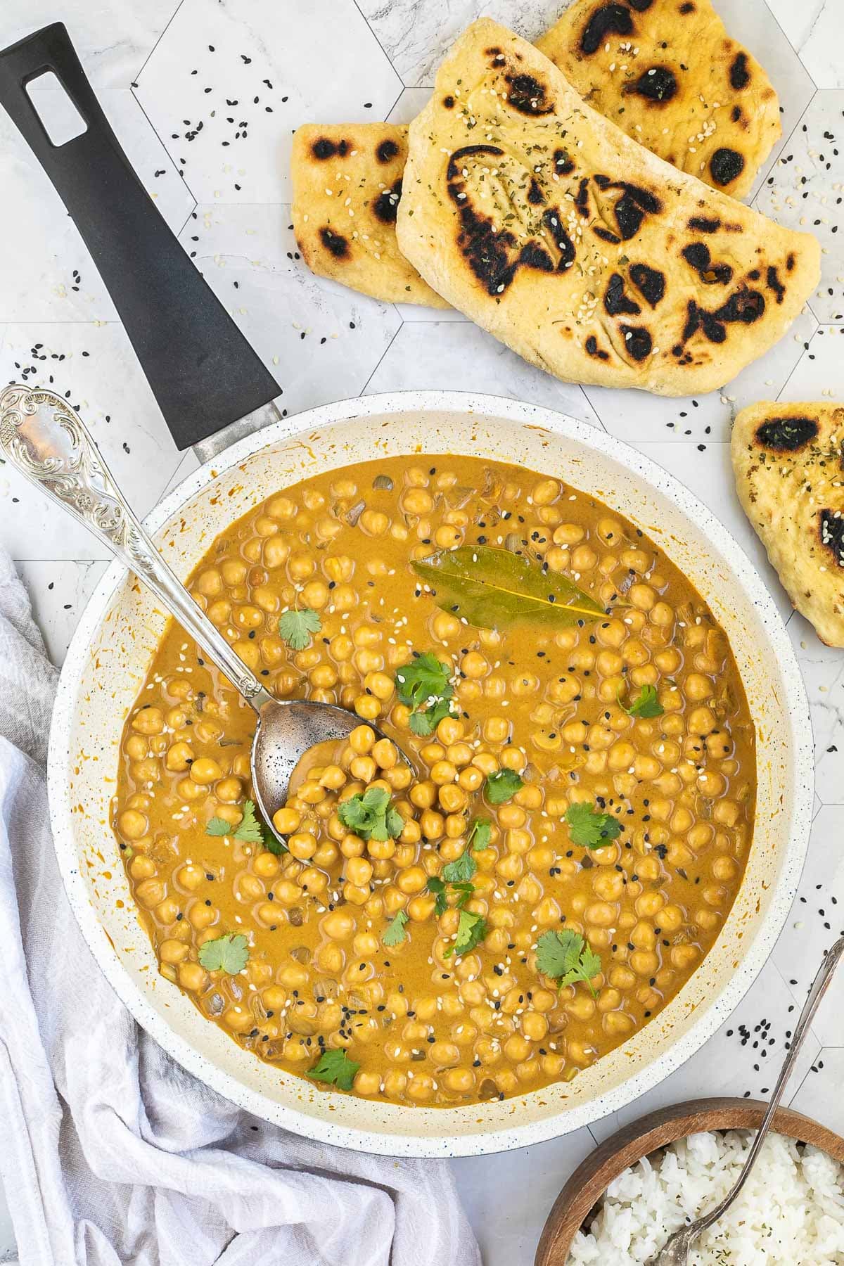 White frying pan with a creamy orange sauce mixed with chickpeas, sprinkled with sesame seeds and freshly chopped cilantro. Charred flatbread and a bowl of rice is next to it.