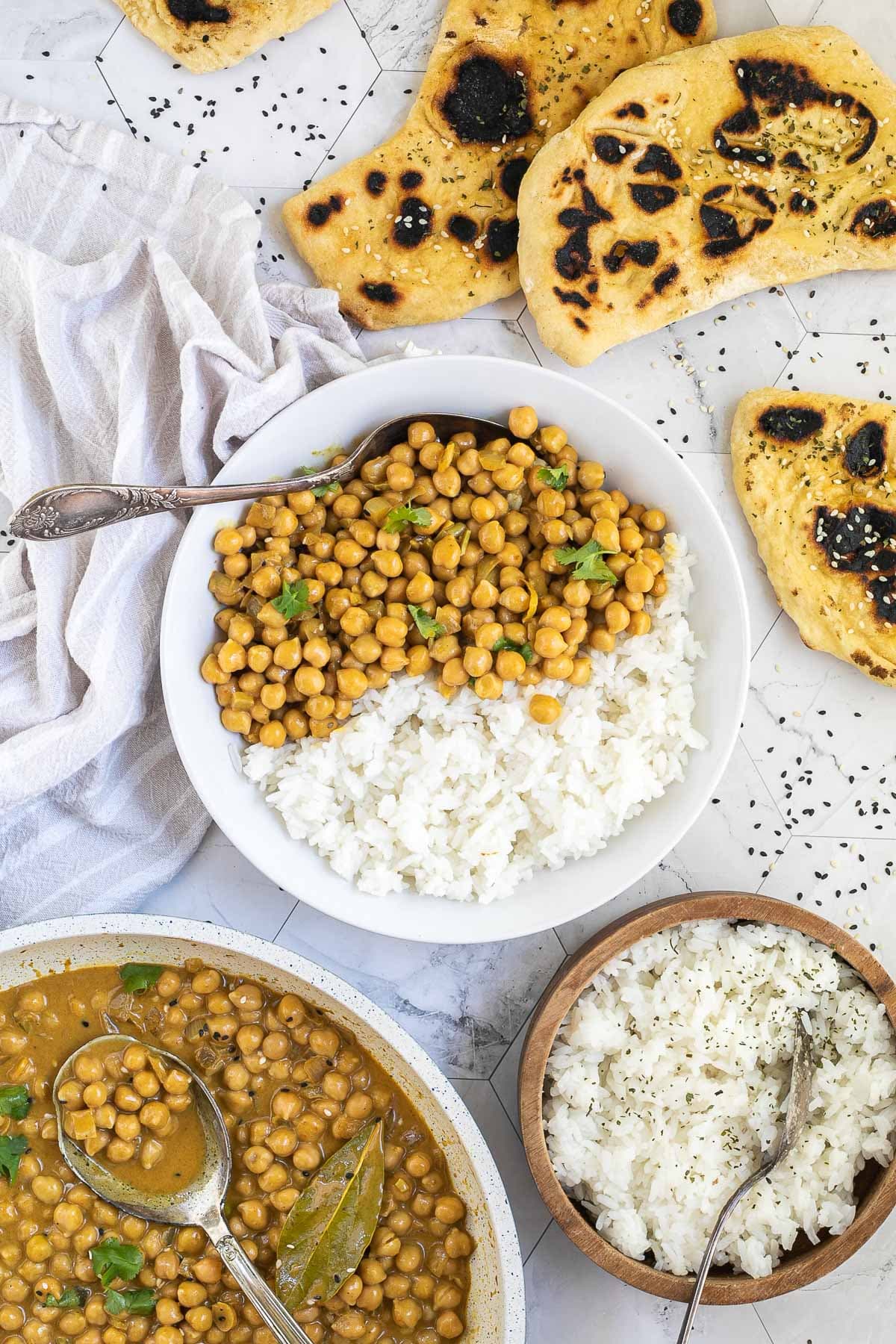 A white plate with white rice and chickpeas in a creamy orange sauce sprinkled freshly chopped cilantro. A spoon is placed inside. Naan bread, a bowl of rice, and leftover chickpea korma are next to it.