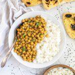 A white plate with white rice and chickpeas in a creamy orange sauce sprinkled freshly chopped cilantro. A spoon is placed inside. Naan bread and a bowl of rice are next to it.