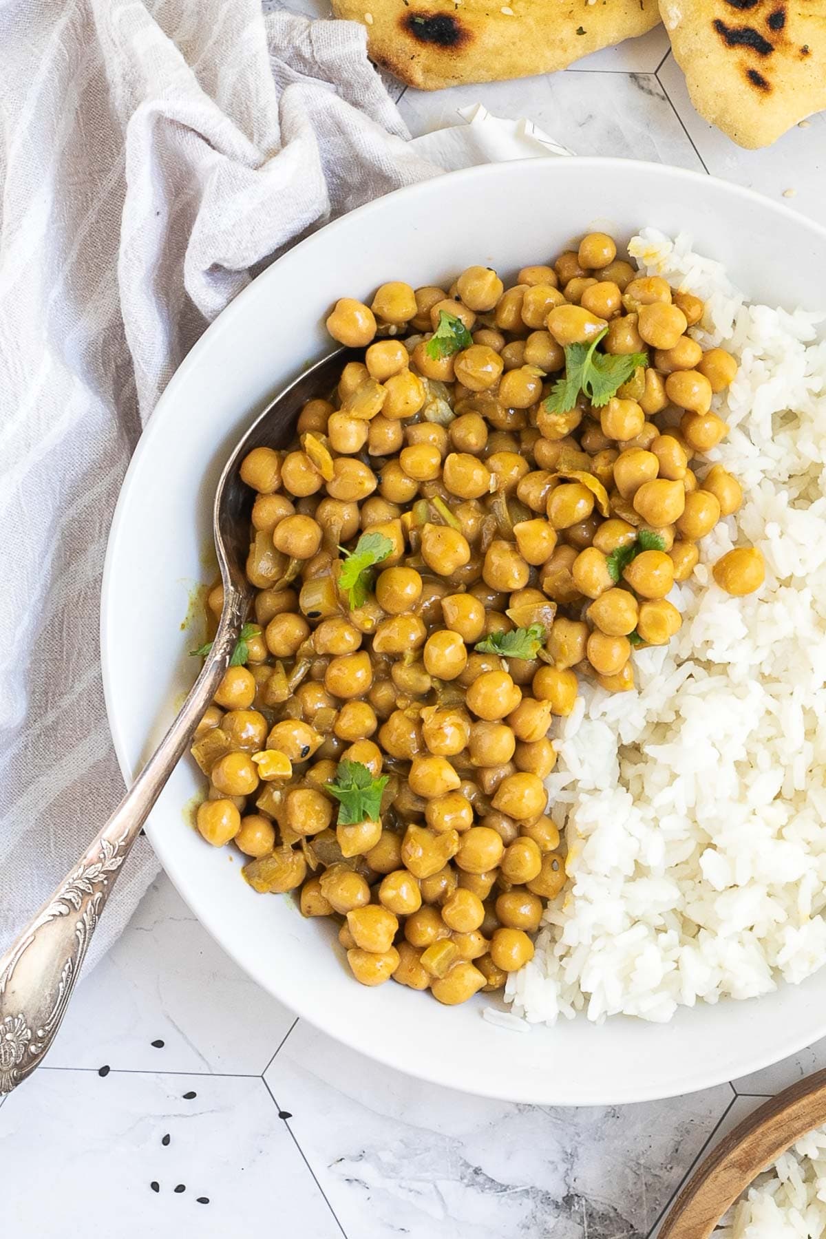 A white plate with white rice and chickpeas in a creamy orange sauce sprinkled freshly chopped cilantro. A spoon is placed inside. Naan bread and a bowl of rice are next to it.