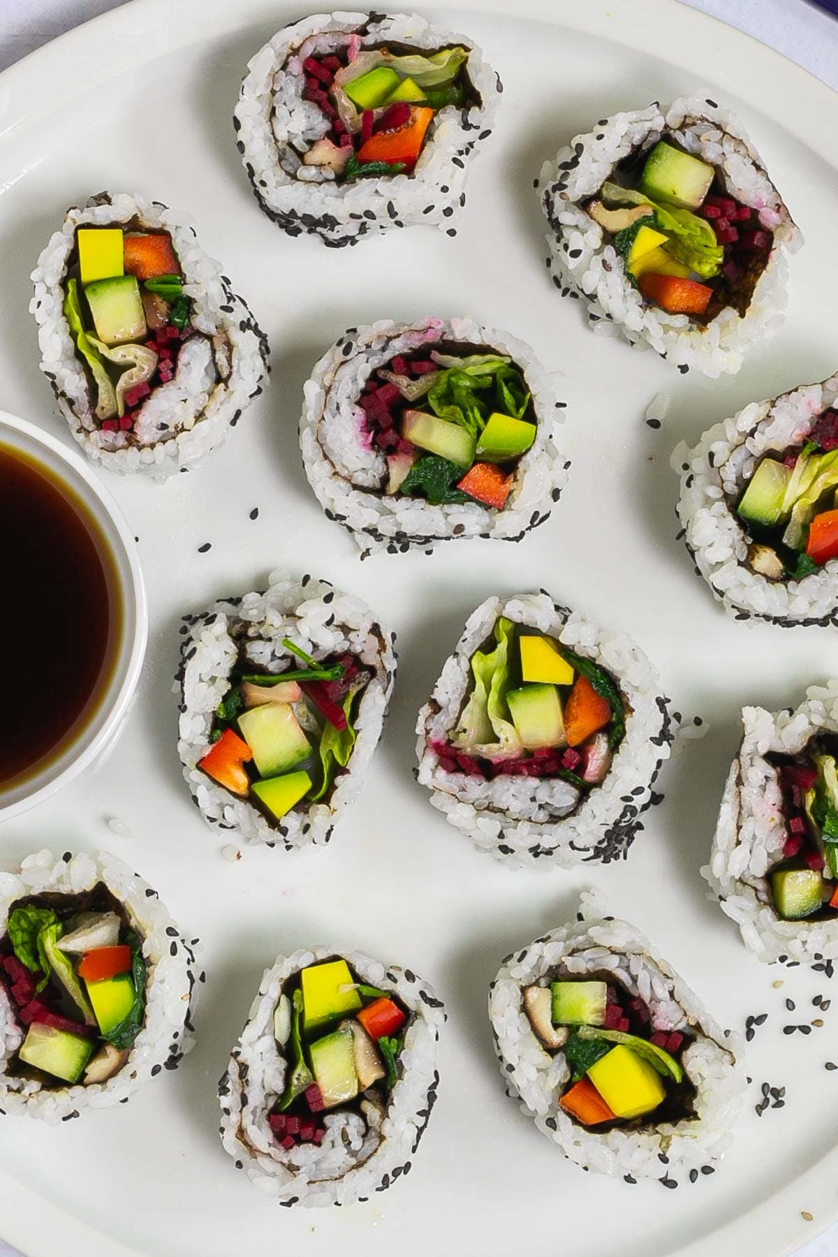 Large white round plate with 11 rainbow sushi rolls without fish or seafood, only vegetables. Served with a small bowl of soy sauce.