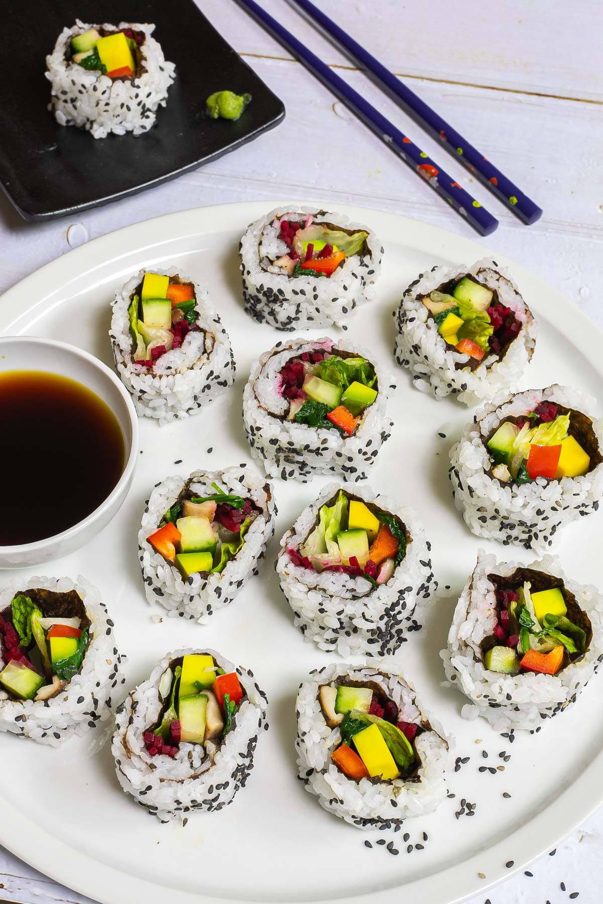 Large white round plate with 11 rainbow sushi rolls without fish or seafood, only vegetables. Served with a small bowl of soy sauce. Blue chopsticks are placed next to it.