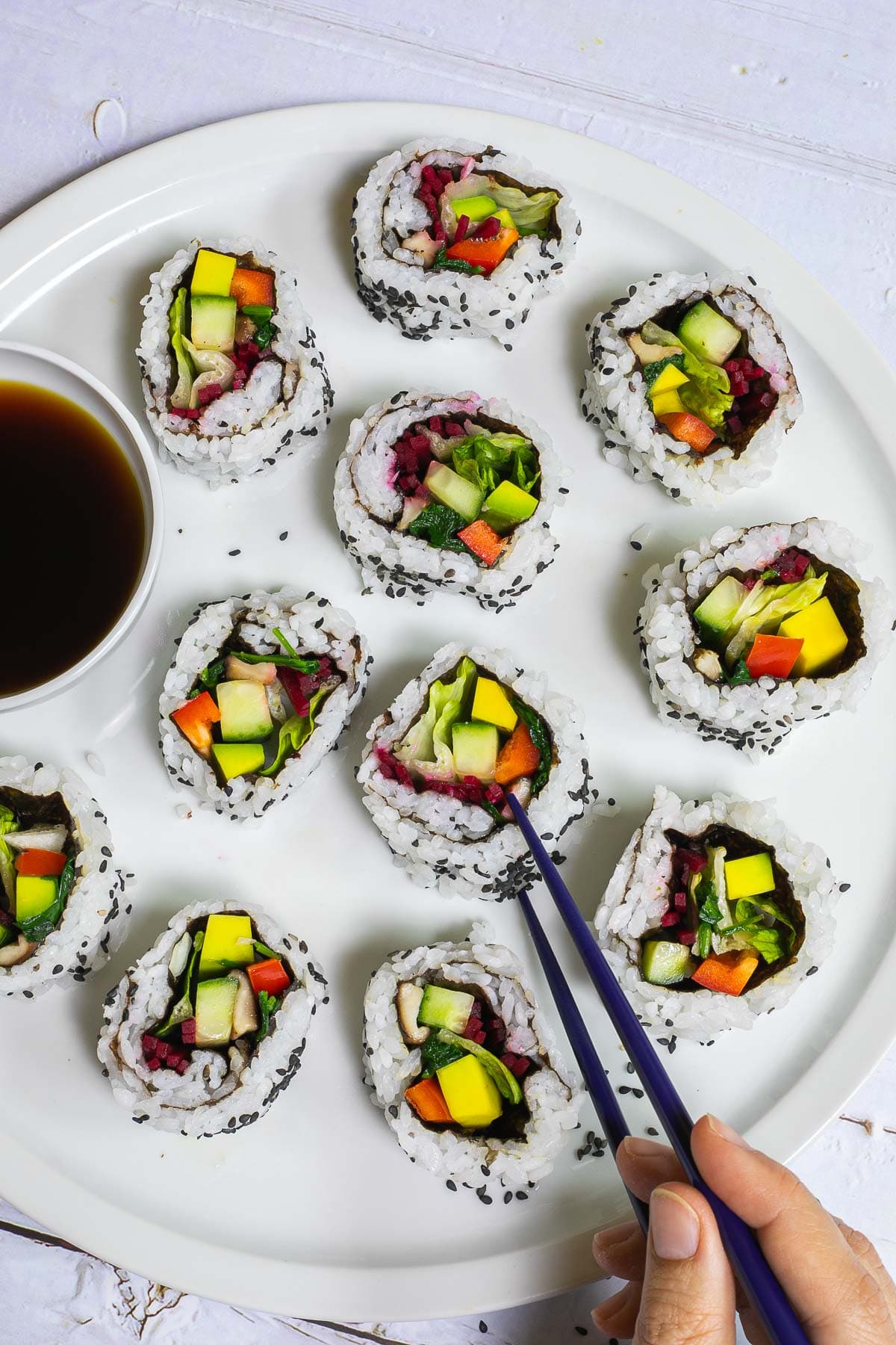 Large white round plate with 11 rainbow sushi rolls without fish or seafood, only vegetables. Served with a small bowl of soy sauce. A hand is holding blue chopsticks to take one.