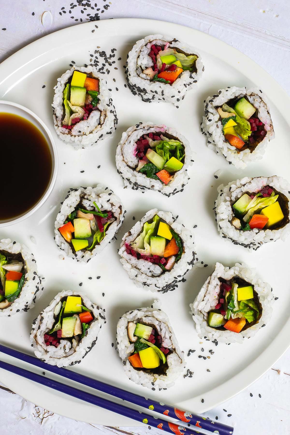 Large white round plate with 11 rainbow sushi rolls without fish or seafood, only vegetables. Served with a small bowl of soy sauce. Blue chopsticks are placed next to it. 