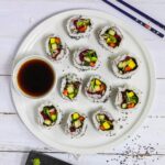 Large white round plate with 11 rainbow sushi rolls without fish or seafood, only vegetables. Served with a small bowl of soy sauce. Blue chopsticks are placed next to it. One sushi is on a black plate separately.