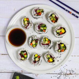 Large white round plate with 11 rainbow sushi rolls without fish or seafood, only vegetables. Served with a small bowl of soy sauce. Blue chopsticks are placed next to it. One sushi is on a black plate separately.