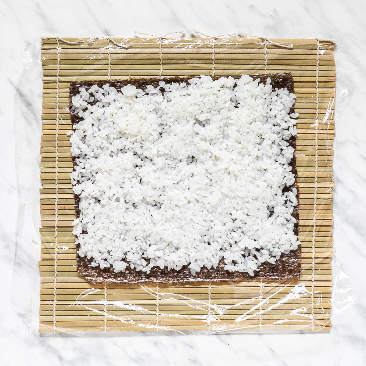 Nori sheets with rice on a bamboo mat covered in plastic wrap.