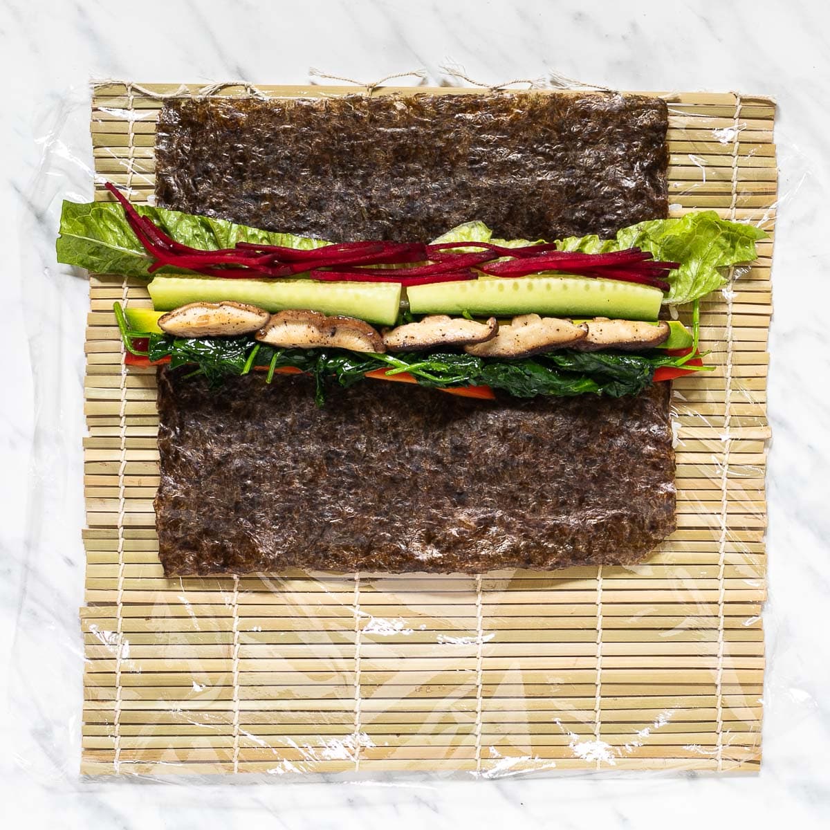 Nori sheets with red pepper, avocado, mushroom, beet, spinach and lettuce slices on a bamboo mat covered in plastic wrap.