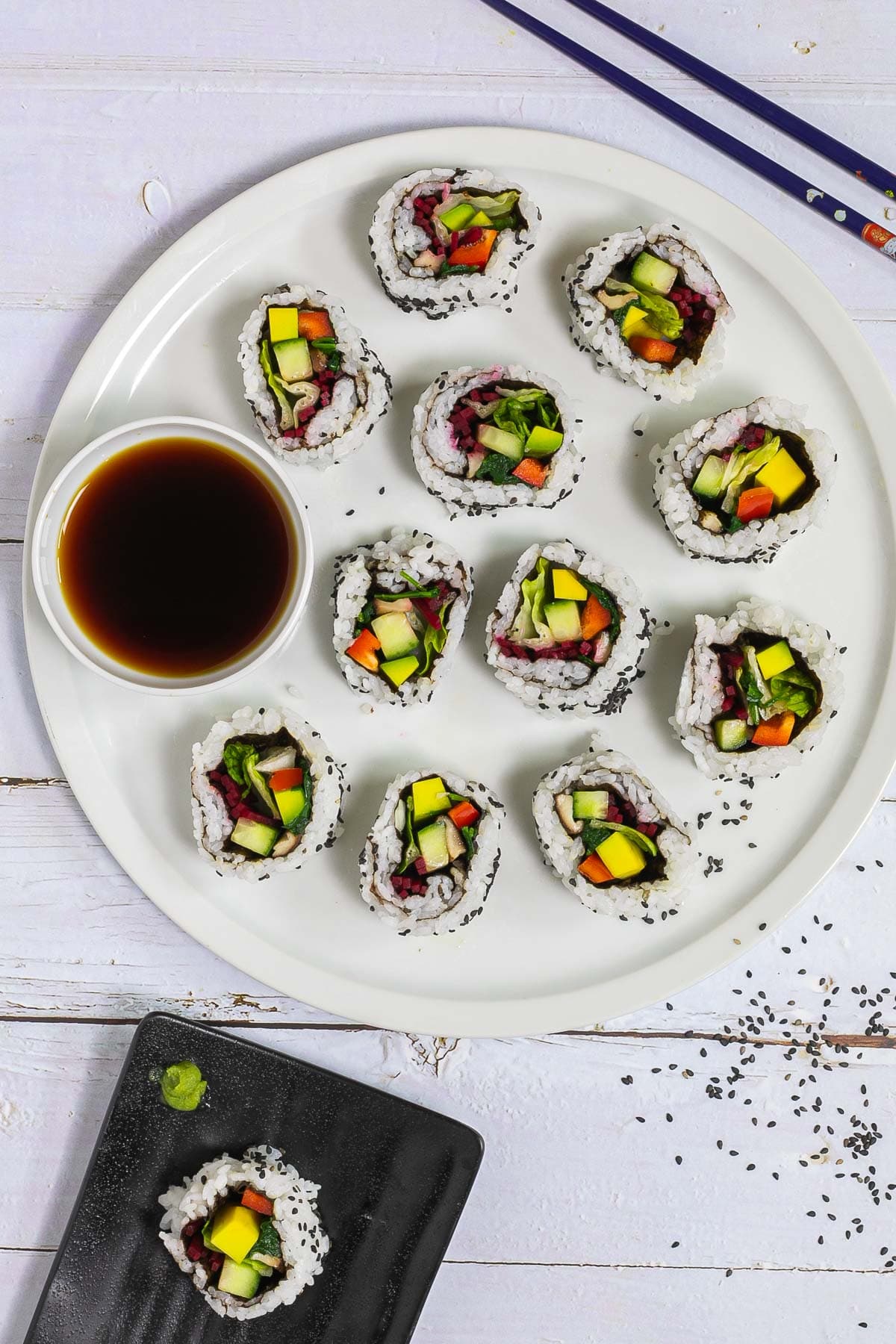 Large white round plate with 11 rainbow sushi rolls without fish or seafood, only vegetables. Served with a small bowl of soy sauce. Blue chopsticks are placed next to it. One sushi is on a black plate separately.