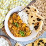 A white plate with white rice, pieces of naan bread and tofu cubes in a creamy orange sauce sprinkled with sesame seeds and freshly chopped cilantro. A spoon is placed inside.