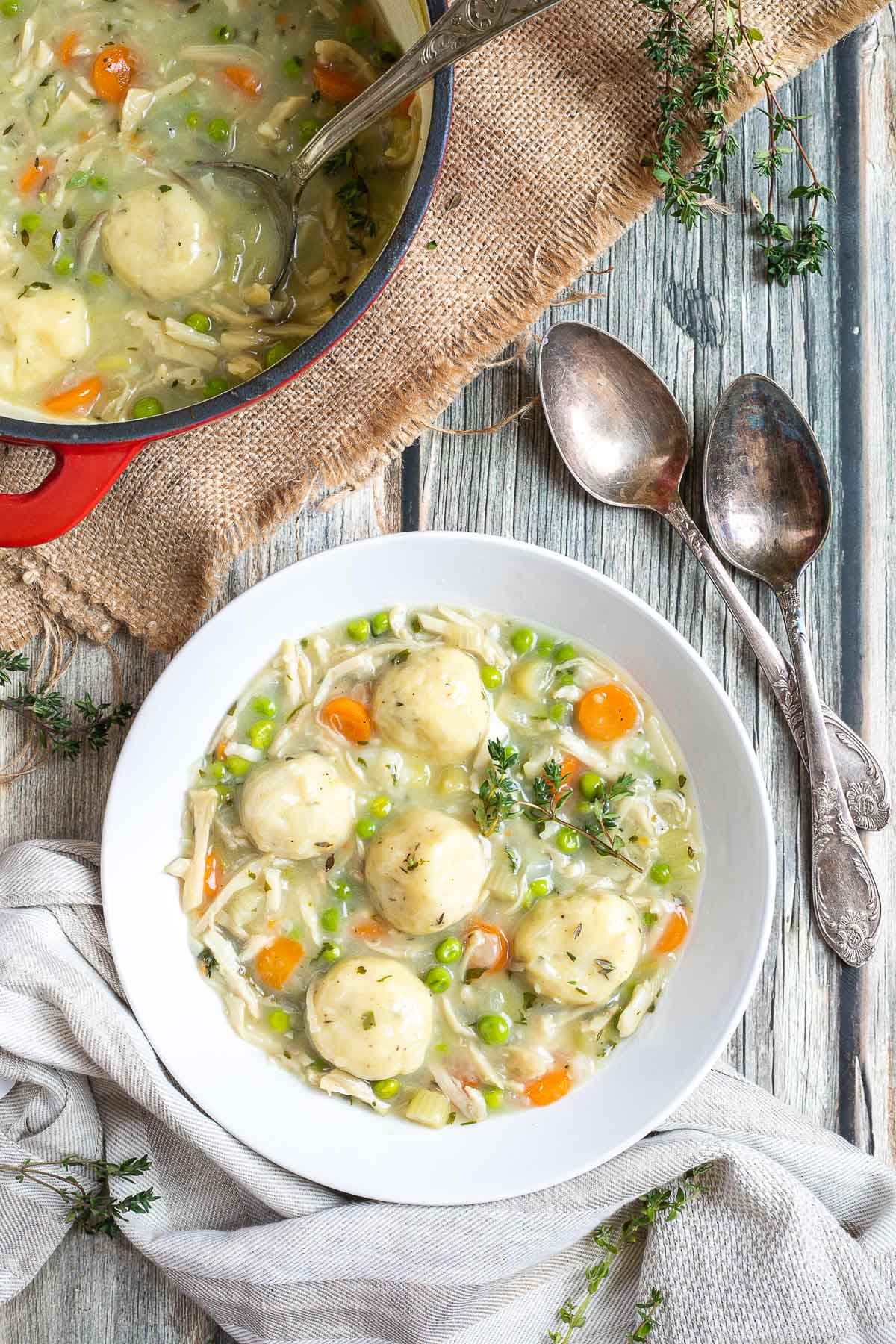 A white plate with dumplings, chopped veggies, shredded mushroom pieces in place of chicken, green herbs in a thick, white stew. The remaining soup is in a red-white enameled Dutch oven next to it.