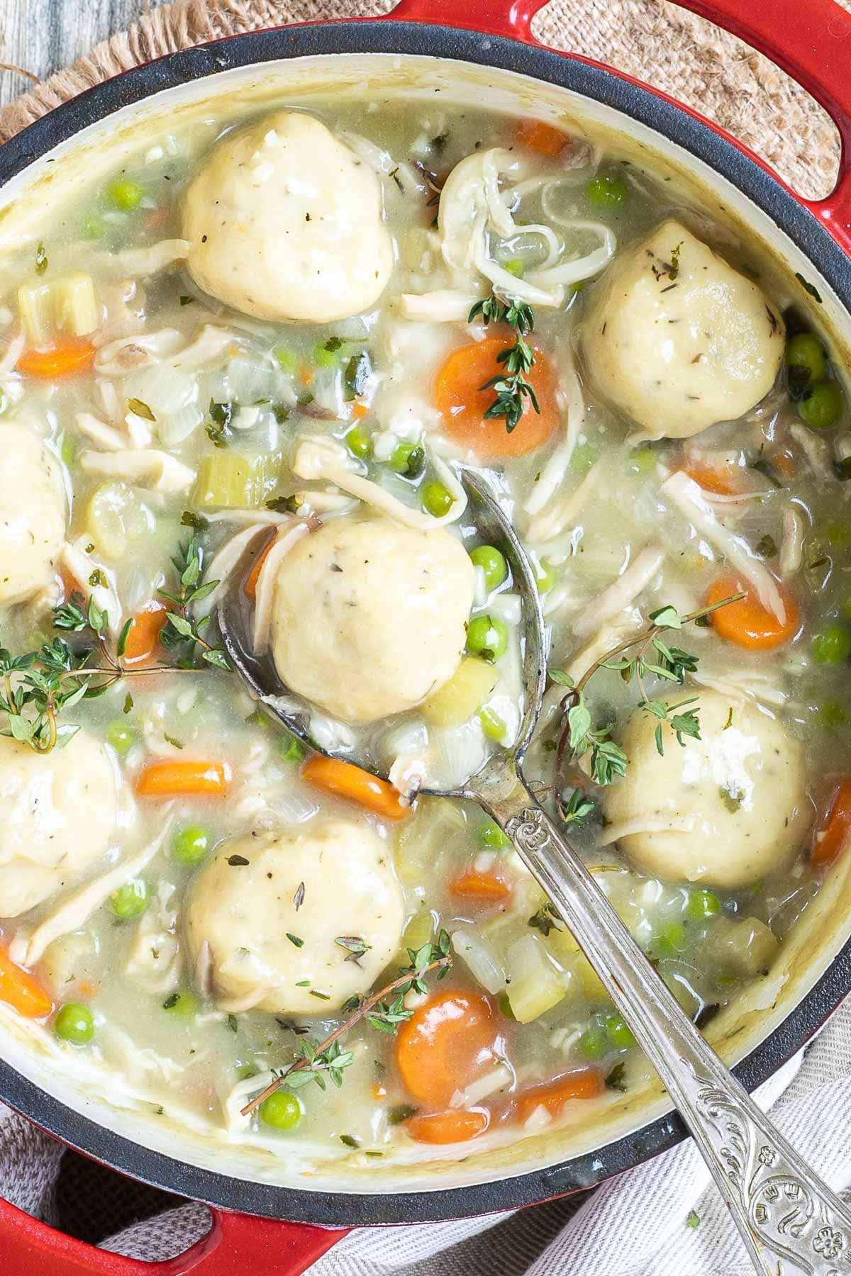 A red-white enameled Dutch oven with dumplings, chopped veggies, shredded mushroom pieces in place of chicken, green herbs in a thick, white stew. A large serving spoon is place in it.