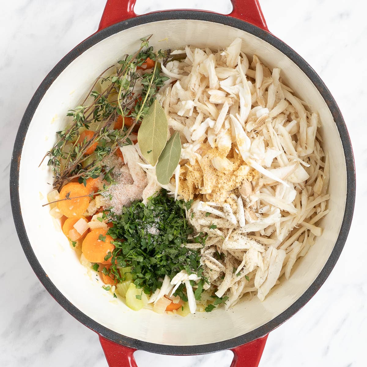 A red-white enameled Dutch oven with chopped veggies, white mushroom shreds, green herbs, and bay leaves.